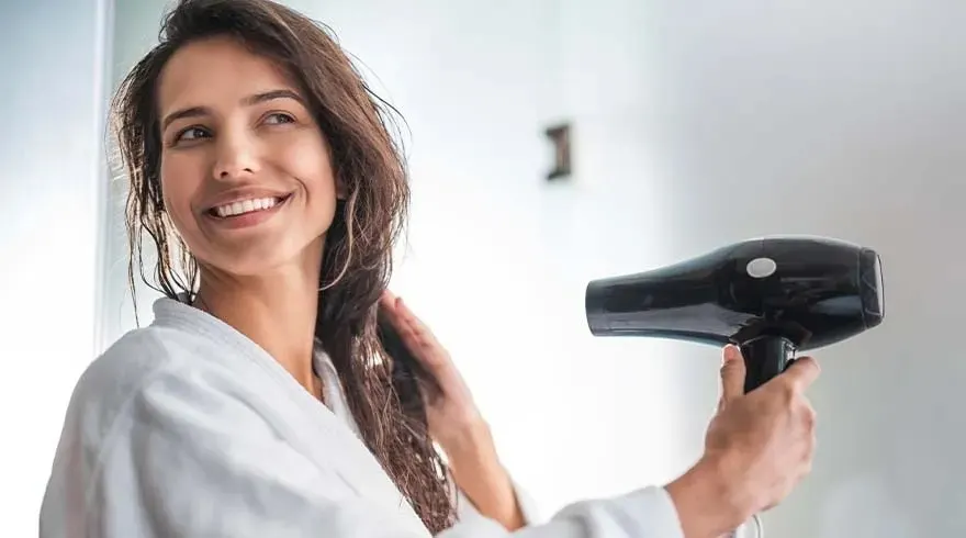 Une salle de bain lumineuse avec un sèche-cheveux noir tenu devant un peignoir blanc, sur fond de mur blanc.
