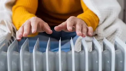 Une personne en pull jaune et sous une couverture blanche touche un radiateur blanc.