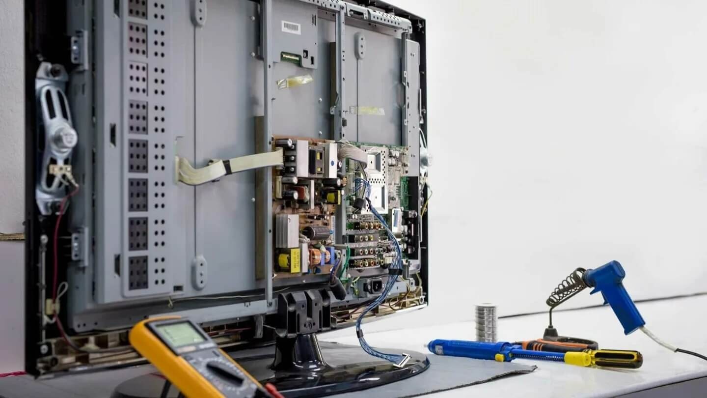 An open television with visible internal electronics, a yellow multimeter, and blue soldering tools on a light surface.