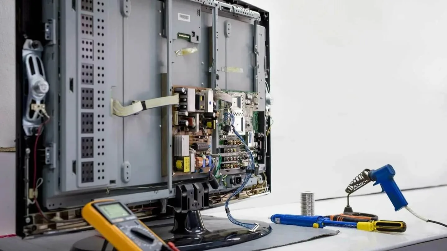 An open television with visible internal electronics, a yellow multimeter, and blue soldering tools on a light surface.