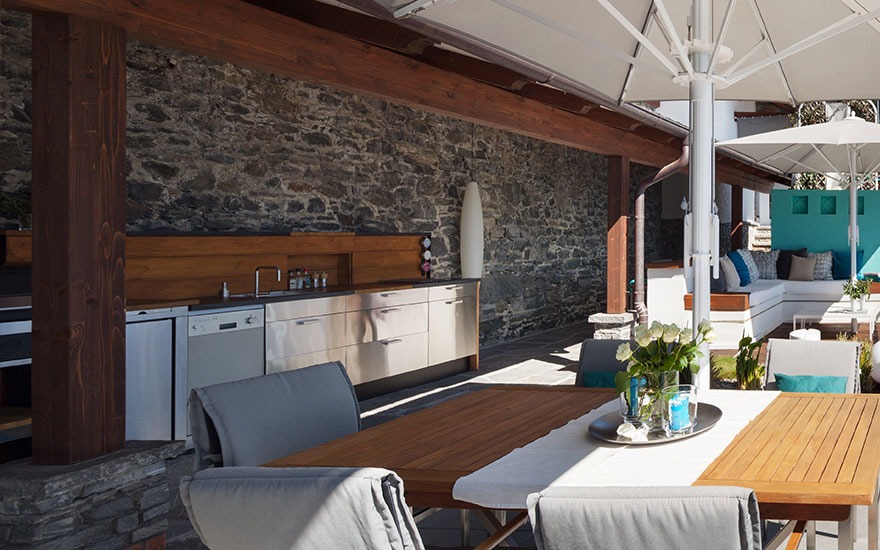 Cuisine extérieure avec mur de pierre, meubles en bois, et parasols blancs. Vaisselle sur table.