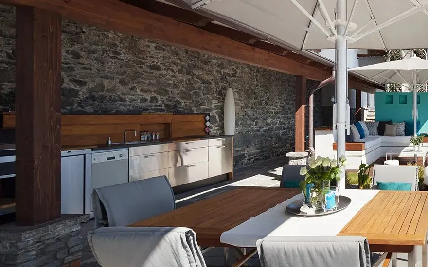 Outdoor patio with stone wall, wooden beams, and kitchen. Tables with chairs, white umbrellas, and blue accents.