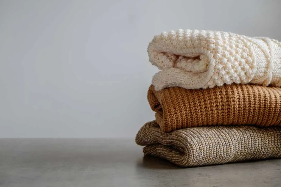 Pile de trois pulls pliés : un crème, un marron et un beige, sur une surface grise, devant un fond gris clair.