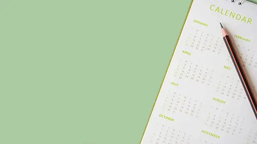 Op een groene achtergrond ligt een kalender met een potlood. De kalender is wit en heeft gele letters en cijfers.