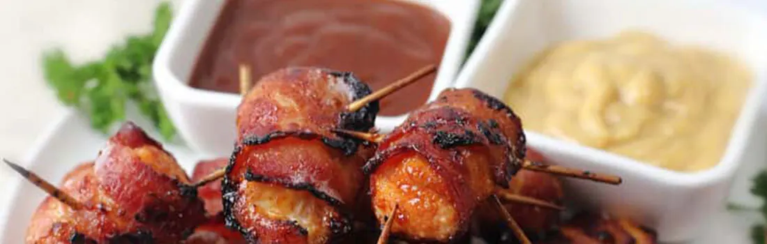 Gros plan sur des brochettes de poulet au bacon avec trempettes brune et jaune, garnies de persil, sur une assiette blanche.