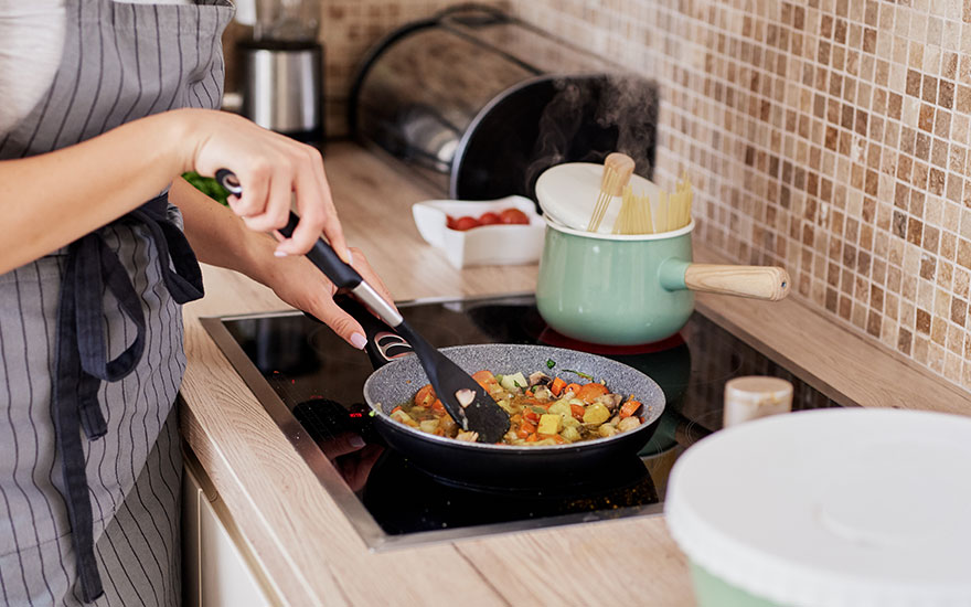 Een keuken met een persoon die groenten roert in een pan op een kookplaat, naast een groene pan.
