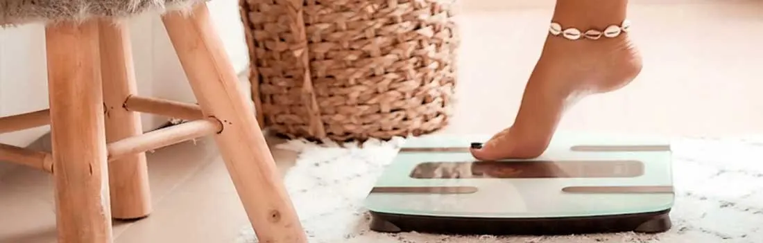 Een persoon stapt blootsvoets op een moderne glazen personenweegschaal. De weegschaal staat op een zacht wit tapijt naast een houten kruk en een gevlochten mand.