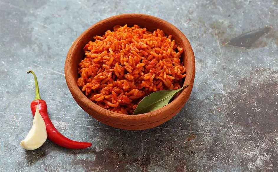 Bol en terre cuite rempli de riz rouge, avec une feuille de laurier. Un piment rouge et une gousse d'ail sont à côté.