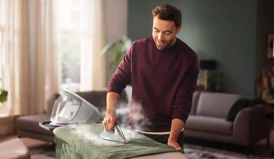 Een stoomstrijkijzer strijkt een groen kleed op een strijkplank, met een stoomgenerator op een zwart platform. Achtergrond met banken.