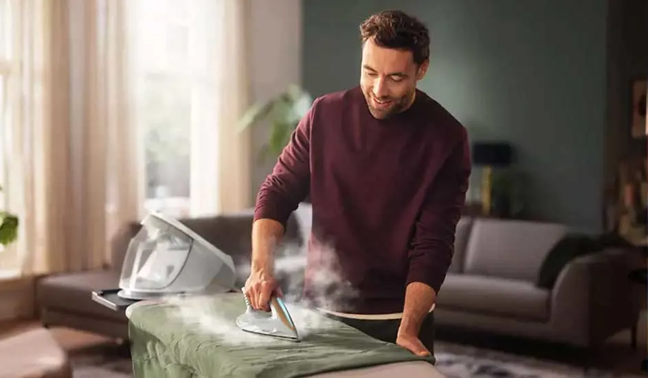 Een stoomstrijkijzer strijkt een groen kleed op een strijkplank, met een stoomgenerator op een zwart platform. Achtergrond met banken.