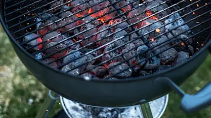 Un barbecue noir avec des briquettes de charbon de bois incandescentes derrière une grille métallique, sur fond d'herbe.