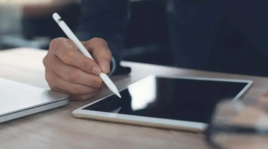 A hand holds a white stylus over a tablet with a black screen on a light brown surface. A white laptop sits to the left.
