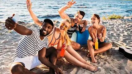 Quatre personnes sur une plage de sable, tenant des boissons près d'une glacière bleue, avec une guitare à proximité. L'océan est en arrière-plan.