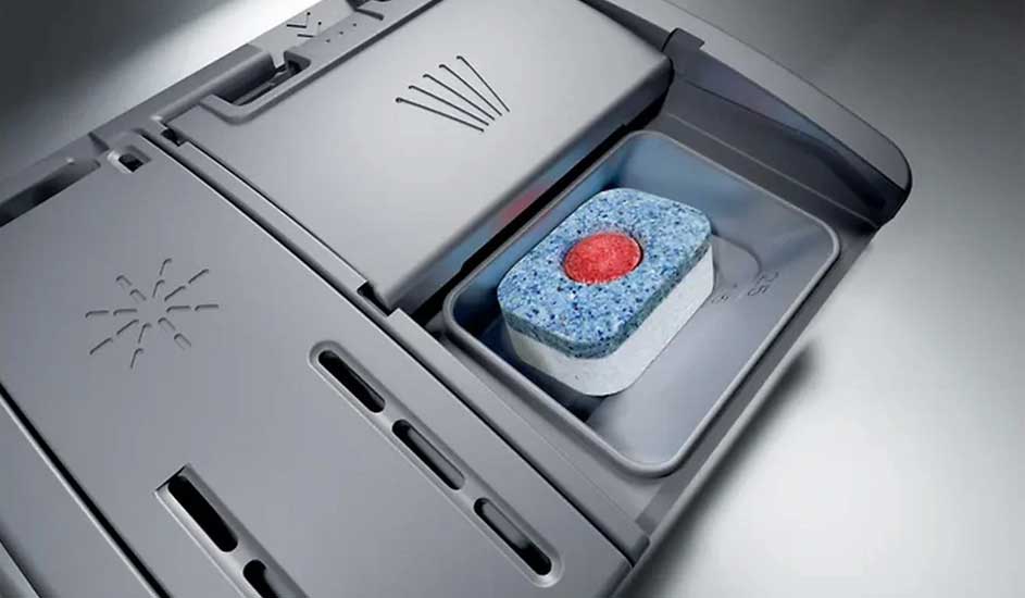 A dishwasher tablet sits in the open detergent compartment of a dishwasher. No brand name is visible on the appliance.