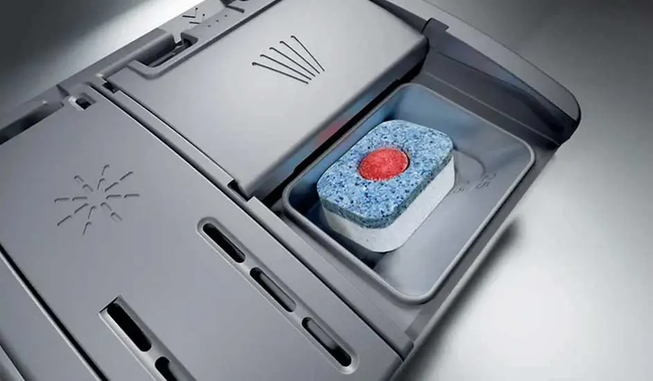 A dishwasher tablet sits in the open detergent compartment of a dishwasher. No brand name is visible on the appliance.