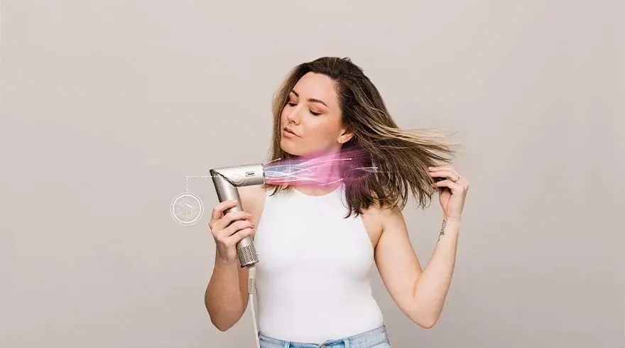 Une femme utilise un sèche-cheveux argenté sur ses cheveux bruns, devant un fond gris uni.