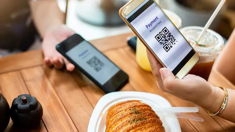 Houten tafel met croissant op wit bord, drankje en twee smartphones met QR-code voor betaling.