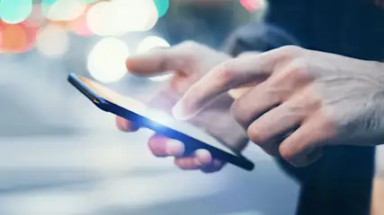 Een hand houdt een zwarte smartphone vast en tikt op het scherm, met gekleurde bokeh-lichten op de achtergrond.