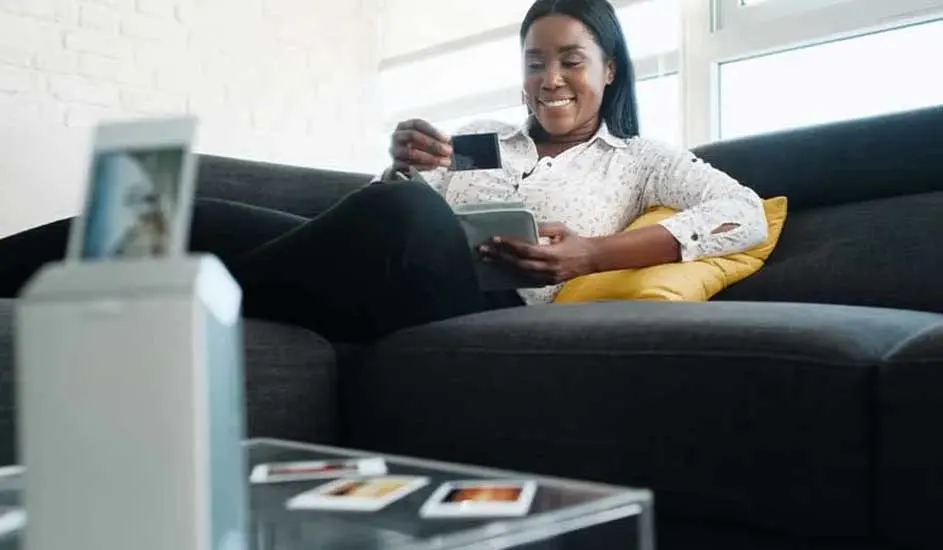 Een zwarte sofa met een geel kussen, een glazen tafel met foto's en een fotoprinter in een lichte kamer.