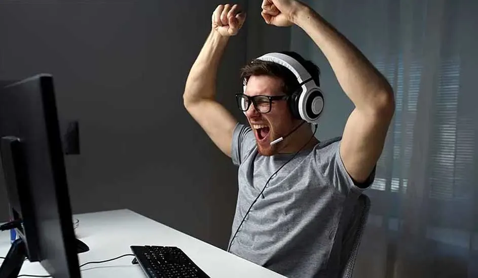 Un écran noir et un clavier sont sur une table blanche. Une personne avec un casque blanc lève les bras. Le fond est gris.