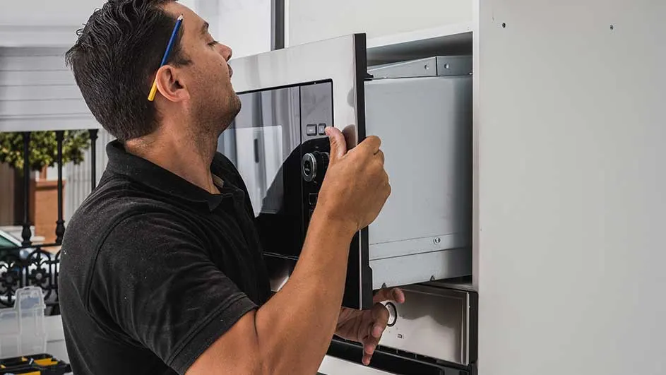 Un homme installe un four à micro-ondes gris encastrable dans une armoire de cuisine blanche.