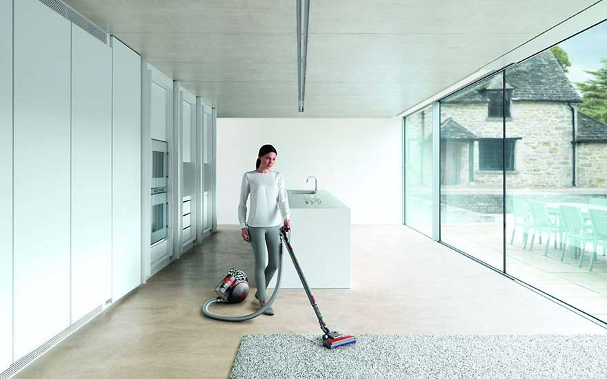 Une pièce moderne avec un aspirateur sur un tapis gris, des armoires blanches et une grande fenêtre donnant sur une maison en pierre.