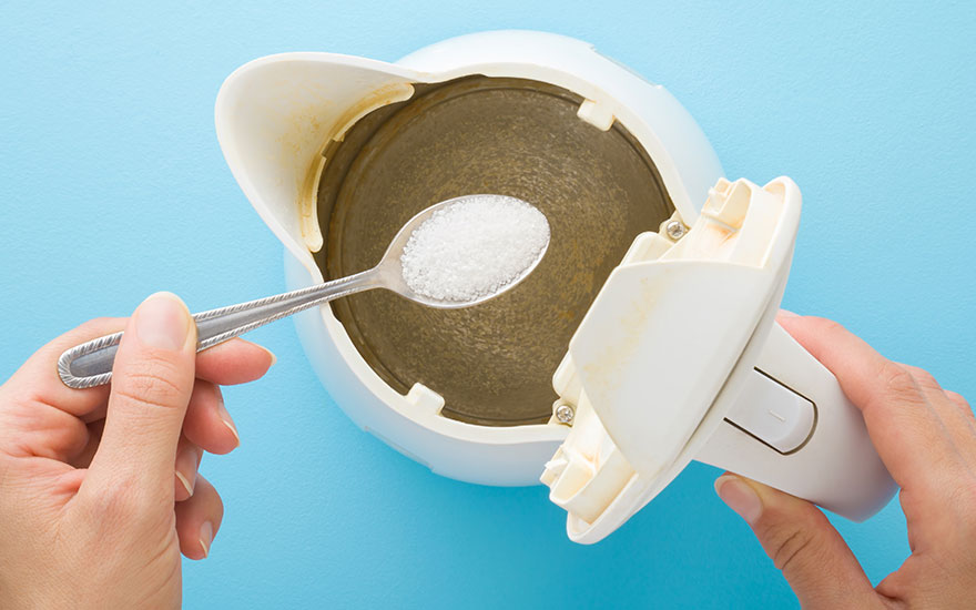 Een witte waterkoker wordt gevuld met een zilveren lepel suiker tegen een blauwe achtergrond.