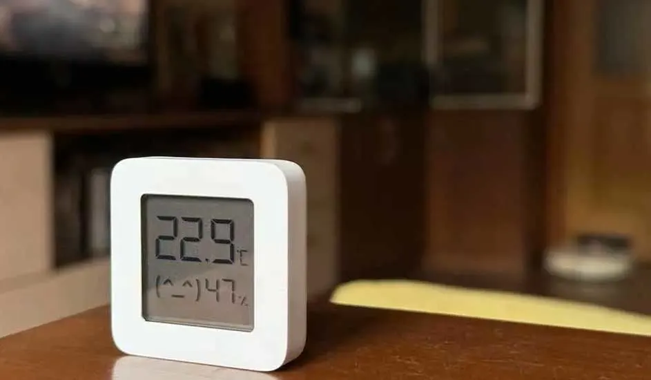 Un thermomètre blanc affiche 22.9 degrés Celsius et une humidité de 47% sur une table marron avec un fond flou.