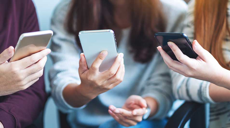 Drie handen houden smartphones vast. De telefoons zijn zwart, zilver en goud. De achtergrond is vaag.