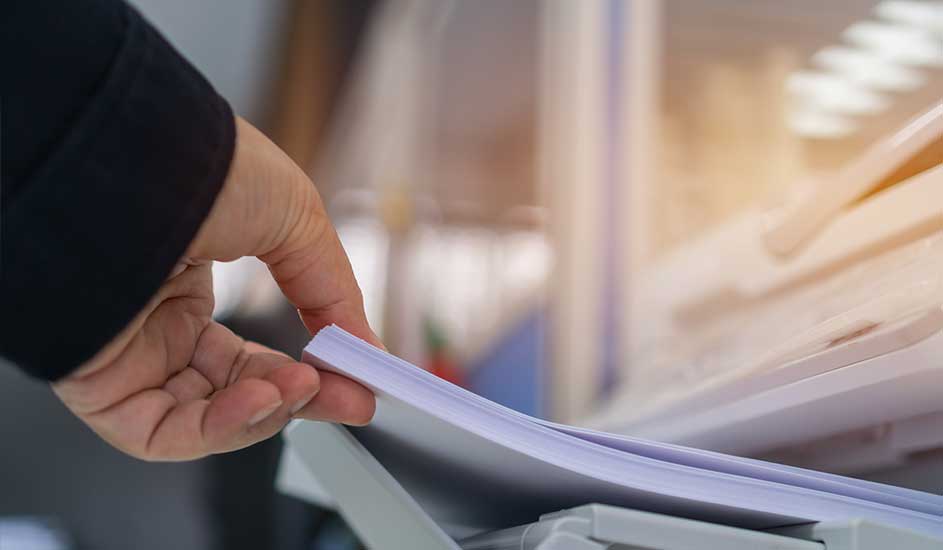 Een hand pakt een stapel wit papier bovenop een grijze printer. Op de achtergrond is een wazig interieur te zien.