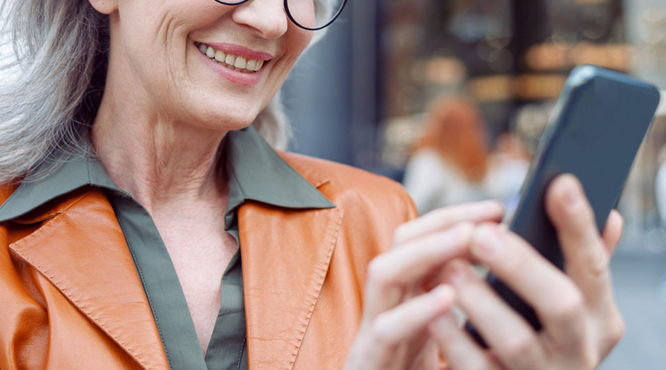 Une personne tient un smartphone. Elle porte une veste orange, une chemise verte et des lunettes. L'arrière-plan est flou.