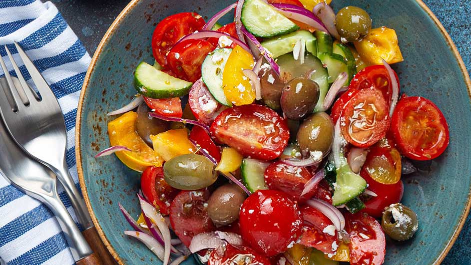 Een blauwe kom gevuld met een salade van tomaten, komkommers, olijven en rode ui. Naast de kom liggen een vork en lepel op een gestreepte doek.