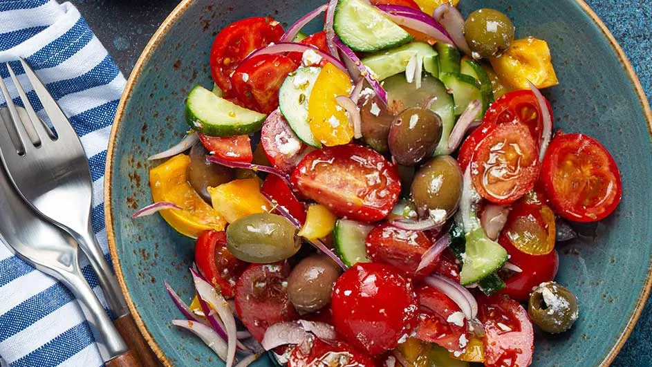 Een blauwe kom gevuld met een salade van tomaten, komkommers, olijven en rode ui. Naast de kom liggen een vork en lepel op een gestreepte doek.