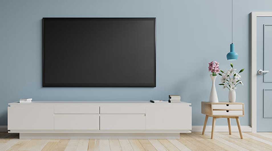 Woonkamer met tv aan een blauwe muur, witte kast, houten vloer, bloemen en blauwe lamp.