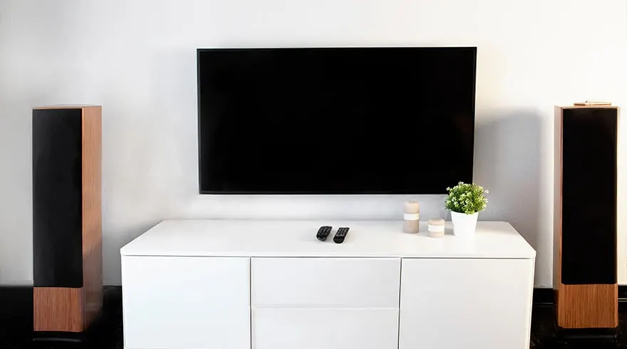 Een woonkamer met een zwarte tv aan een witte muur, witte kast, twee luidsprekers, plant en afstandsbedieningen.