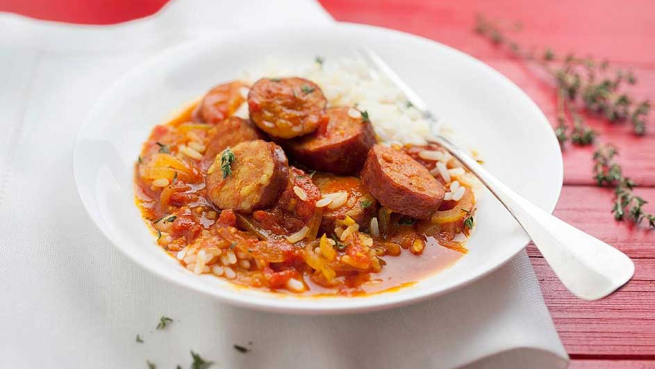 Assiette blanche avec riz, saucisses et sauce tomate, fourchette à droite, sur fond rouge et tissu blanc.