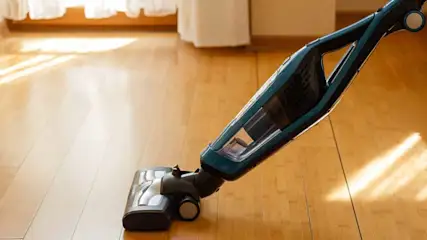 Un aspirateur bleu et noir sur un plancher en bois clair, près d'une fenêtre avec des rideaux blancs.