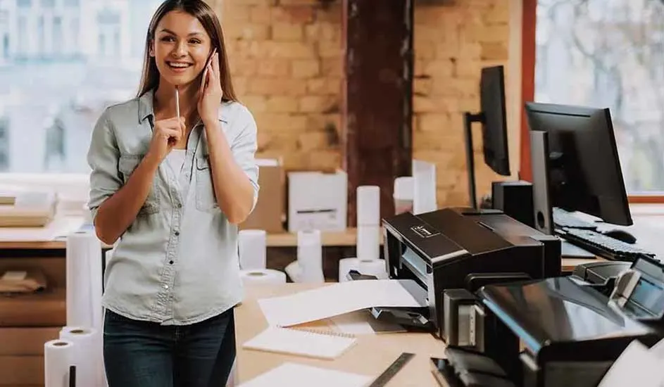 Een bureau met een printer, papier, computerschermen en kantoorbenodigdheden tegen een bakstenen achtergrond.