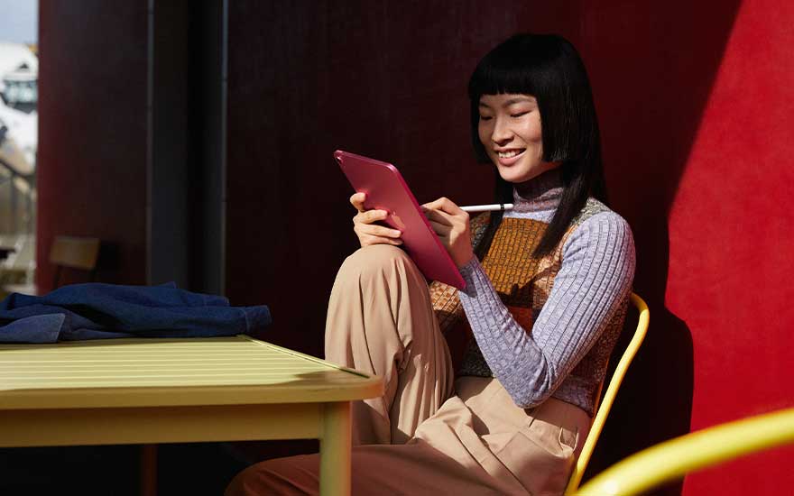 Une tablette rose est utilisée avec un stylet devant un mur rouge, à côté d'une table jaune avec un vêtement bleu.