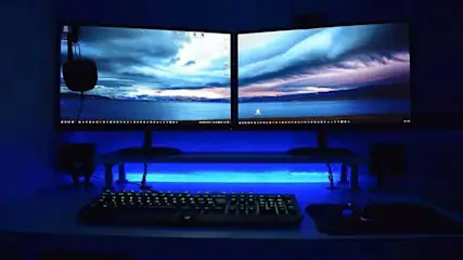Twee monitoren tonen een landschap met blauwe zee en bewolkte lucht. Een toetsenbord en muis staan op een bureau met blauwe verlichting.