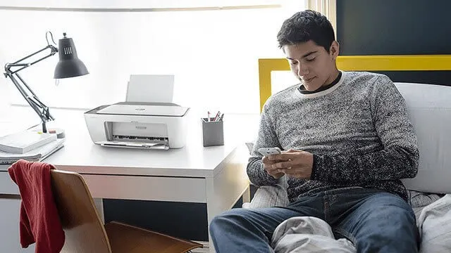 Un bureau blanc avec une imprimante, une lampe et des livres. Une chaise rouge est devant. Un lit avec une tête de lit jaune est à côté.