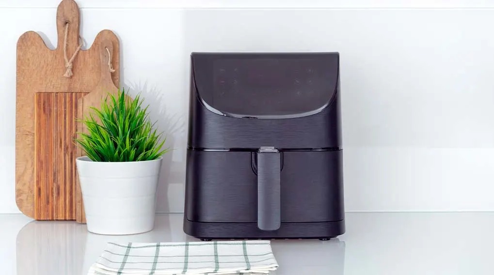 Une friteuse noire est sur un comptoir blanc, à côté d'une plante en pot et de planches à découper en bois. Une serviette à carreaux est au premier plan.