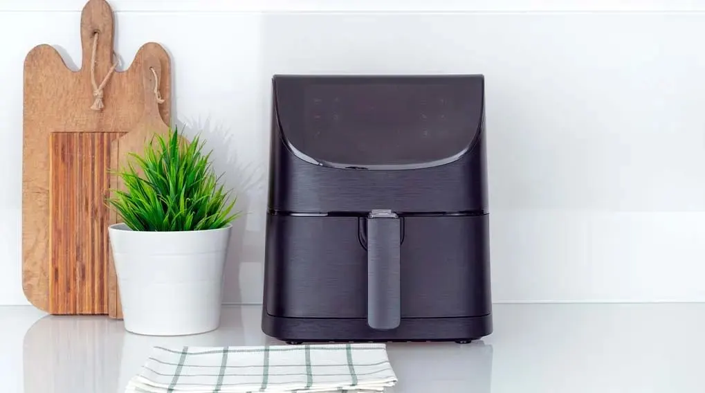 Une friteuse noire est sur un comptoir blanc, à côté d'une plante en pot et de planches à découper en bois. Une serviette à carreaux est au premier plan.