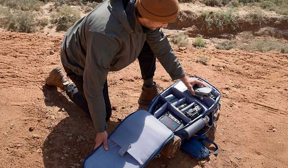 Un homme est à genoux sur le sable et sort un drone d’un sac photo bien rempli, avec divers compartiments et équipements. Aucune marque visible.