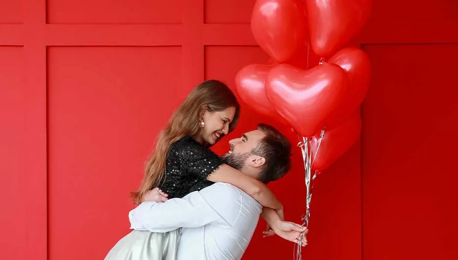 A loving couple against a bright red wall; he lifts her in his arms as they smile at each other beside a cluster of red heart balloons.