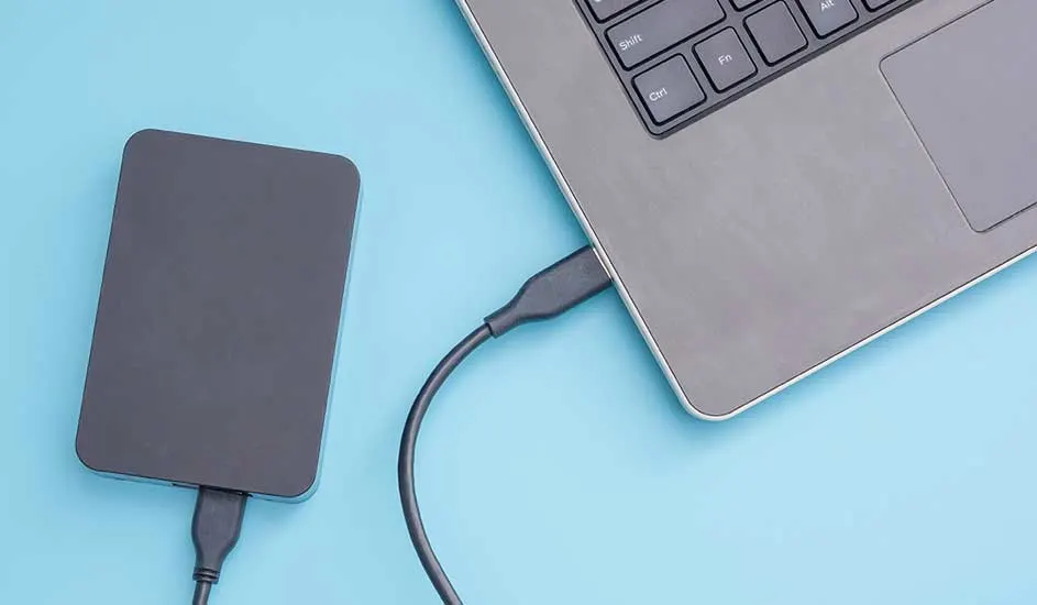 Un disque dur noir est connecté à un ordinateur portable gris sur un fond bleu clair.