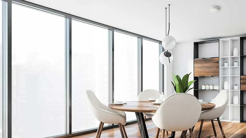 Eetkamer met ronde tafel, witte stoelen en grote ramen. Een plant en een lamp hangen boven de tafel. Een witte kast staat achteraan.