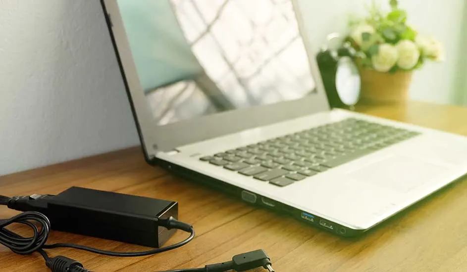 Un ordinateur portable gris est posé sur une table en bois, avec un chargeur noir et une plante floue en arrière-plan.