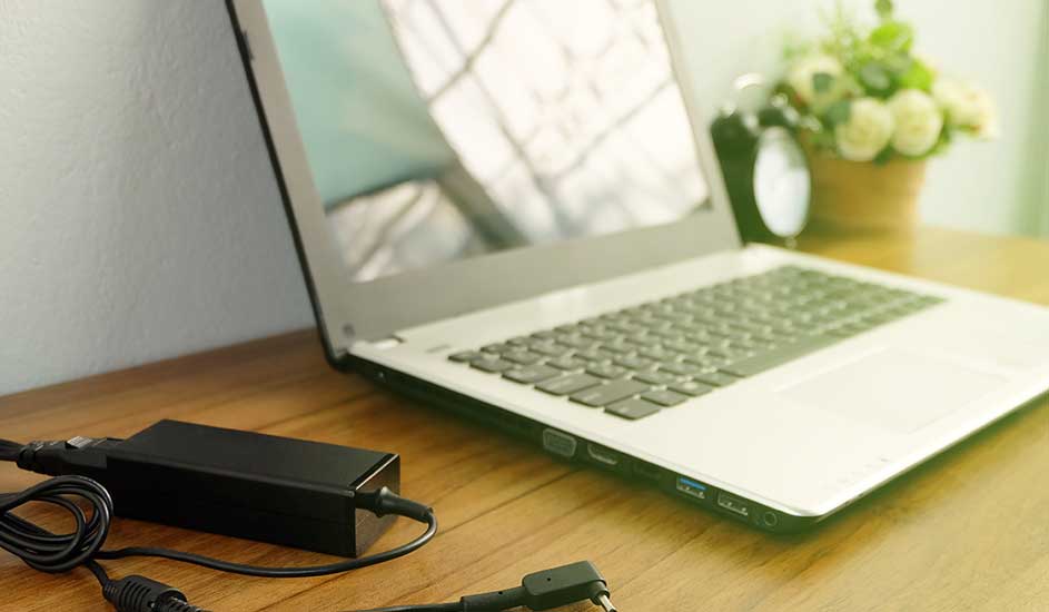 Een open laptop met een zwarte adapter op een houten bureau. Een bloempot staat op de achtergrond.