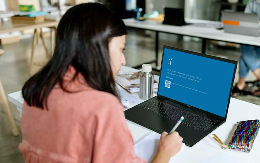Bureau blanc avec ordinateur portable affichant un écran bleu d'erreur, une bouteille d'eau, un cahier et un étui coloré.